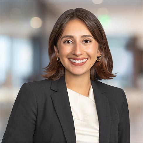 A woman with short brown hair, wearing a black blazer and white top, smiles at the camera in a modern corporate law office with a blurred background.