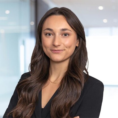 A woman with long brown hair wearing a black blazer stands in a modern corporate law office with glass walls and bright lighting.