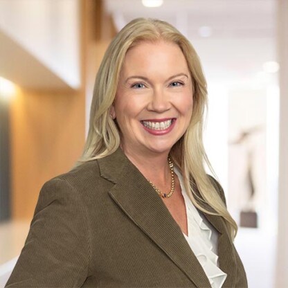 A woman with long blonde hair, wearing a brown blazer, white blouse, and gold necklace, smiles while standing in a brightly lit law office hallway specializing in intellectual property law.