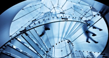 View from below of a modern glass spiral staircase in a corporate law office, with blurred silhouettes of people walking above, illuminated by blue-toned light.