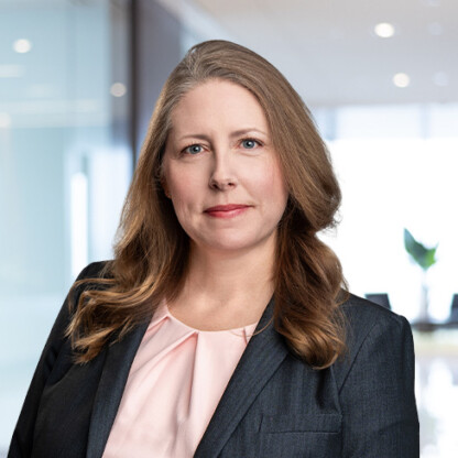 Woman with long light brown hair wearing a dark blazer and pink blouse, standing in a modern law office with glass walls and blurred background—reflecting the professional environment of top lawyers in Chicago.
