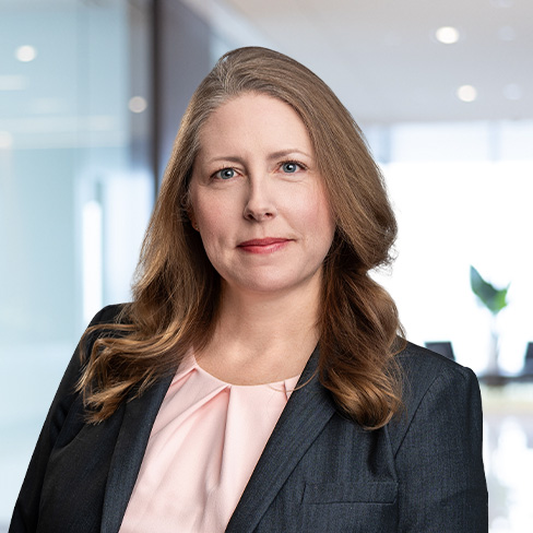 Woman with long light brown hair wearing a dark blazer and pink blouse, standing in a modern law office with glass walls and blurred background—reflecting the professional environment of top lawyers in Chicago.