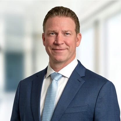 A man in a blue suit and light blue tie stands in front of a blurred corporate law office background, facing the camera and smiling slightly.