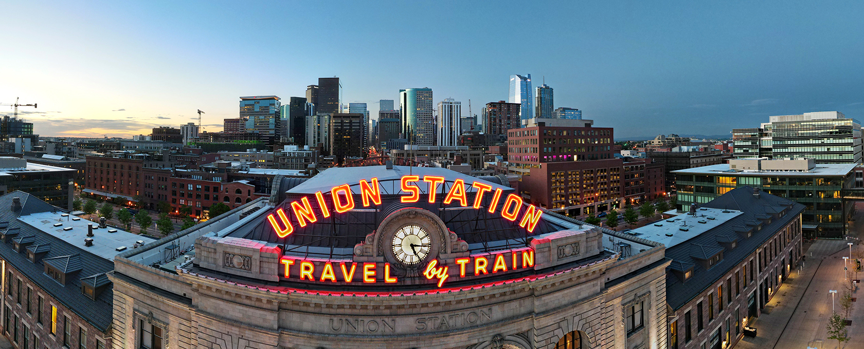 Union Station building with illuminated "Travel by Train" sign in front, neighboring a corporate law office, set against a city skyline at dusk.