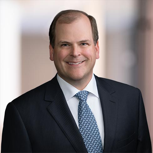 A man in a dark pinstripe suit, white shirt, and patterned tie is smiling at the camera against a blurred indoor background of a corporate law office.