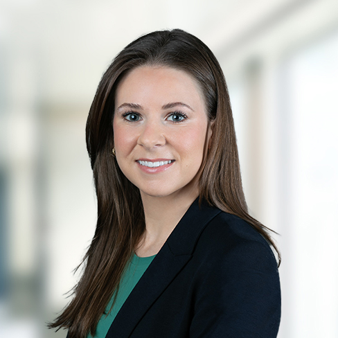 A woman with straight brown hair, wearing a dark blazer over a green top, is smiling at the camera in a bright, blurred indoor setting—representing accomplished Chicago lawyers.