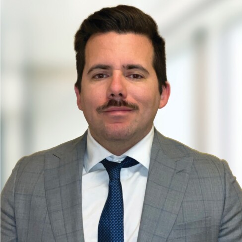 A man with dark hair and a mustache, dressed in a gray suit, white shirt, and blue tie, stands in front of a blurred indoor background—reflecting the professional image of top lawyers in Chicago.