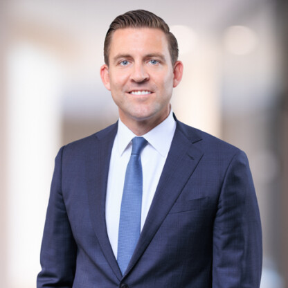 Man in a navy blue suit and tie standing in front of a blurred corporate law office background, facing the camera and smiling.