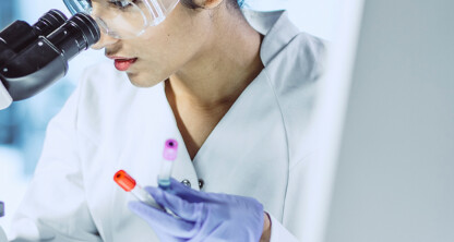 A scientist wearing protective glasses examines samples under a microscope while holding two test tubes in a laboratory, providing crucial research that can aid intellectual property law and litigation support for lawyers in Chicago.