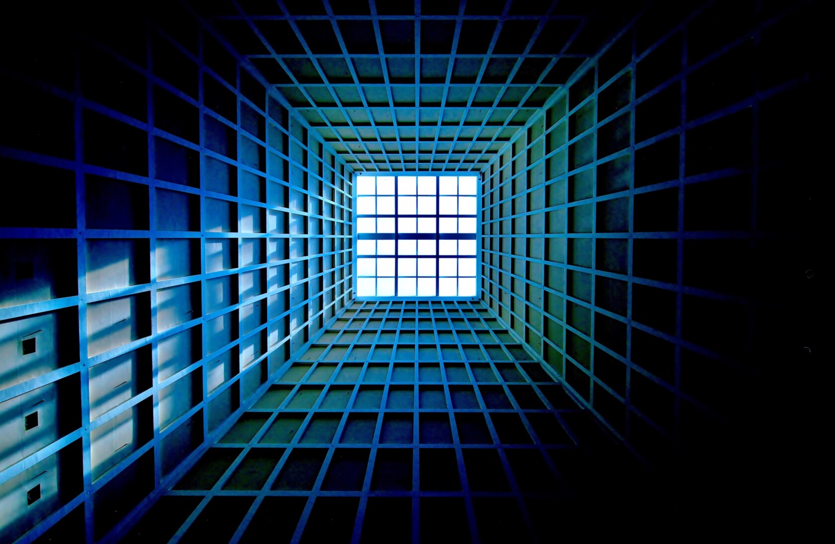 A geometric grid structure viewed from below, with blue and green tones, leading towards a bright white square opening at the top center.