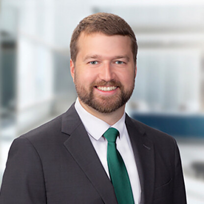 A man with short brown hair and a beard, wearing a dark suit, white shirt, and green tie, smiles at the camera in a modern office setting.