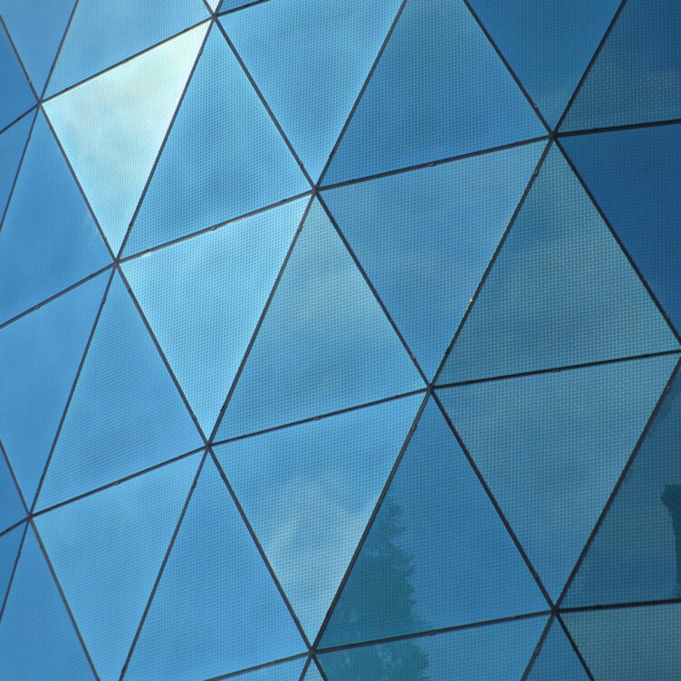 Glass building facade with triangular panels reflecting blue sky and clouds.