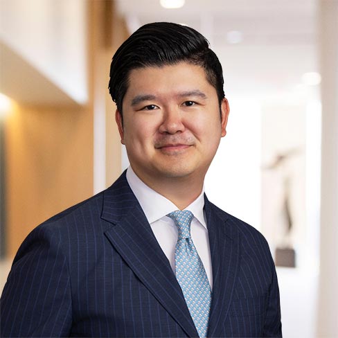 A man in a blue pinstripe suit and light blue tie stands in a well-lit hallway, facing the camera and smiling slightly.