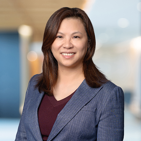 A woman with straight brown hair, wearing a blue blazer over a maroon top, smiles at the camera with a blurred office background.