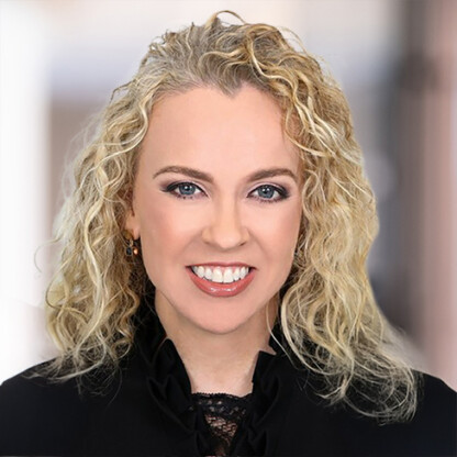 A woman with curly blonde hair, light skin, and blue eyes is smiling at the camera, wearing a black top. The background is softly blurred.