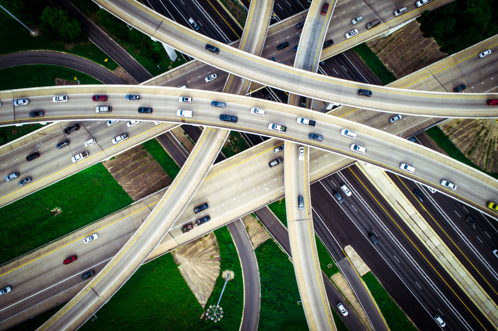 Aerial view of a busy multilayered highway interchange with cars traveling in multiple directions, surrounded by patches of green grass and brown fields.